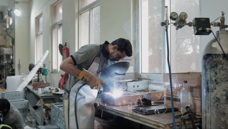 A welder in gloves and apron welding a steel enclosure at a bench